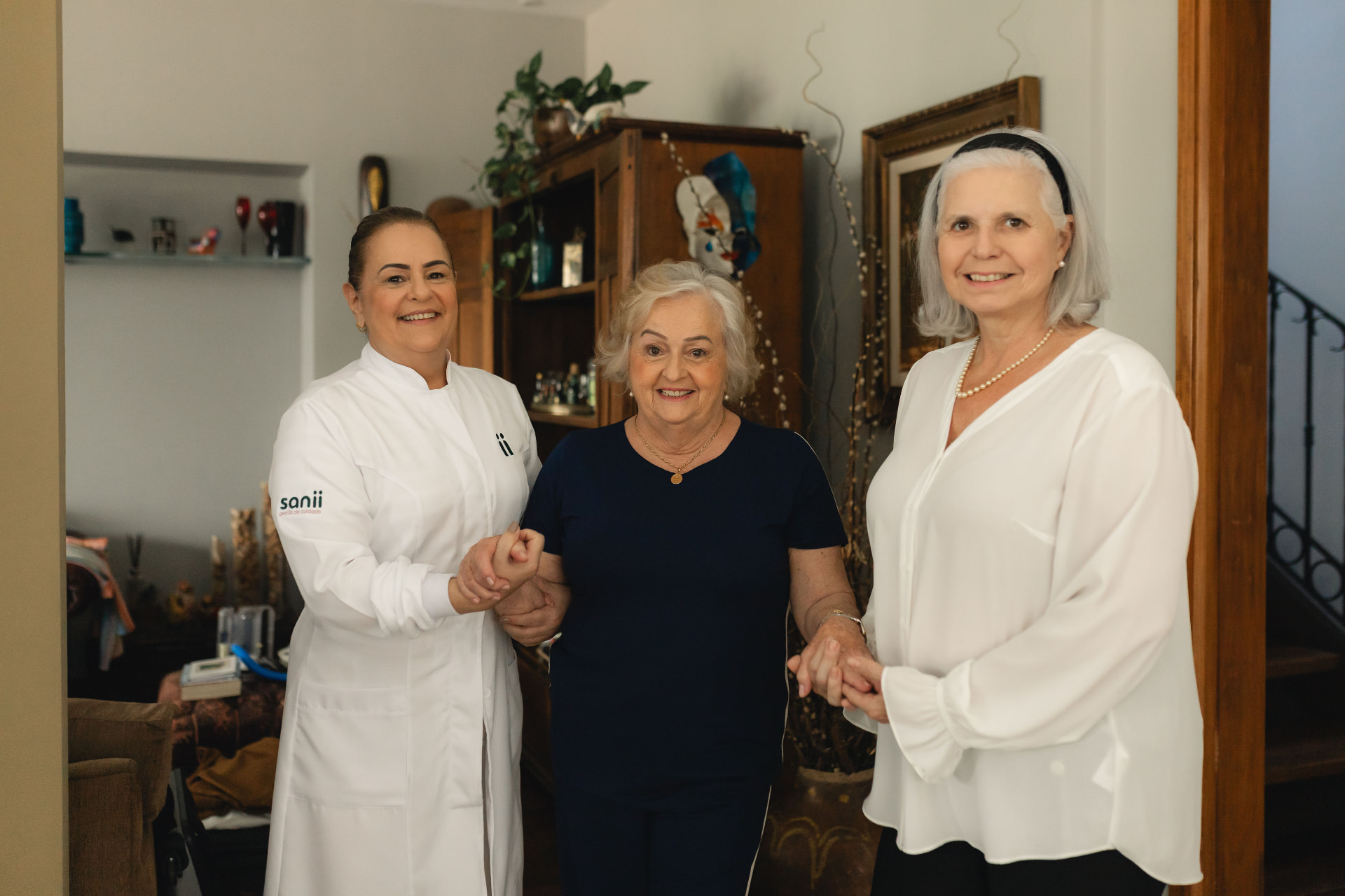 Cuidadora da Sanii ao lado de uma senhora idosa e de sua filha, todas sorrindo na sala de casa
