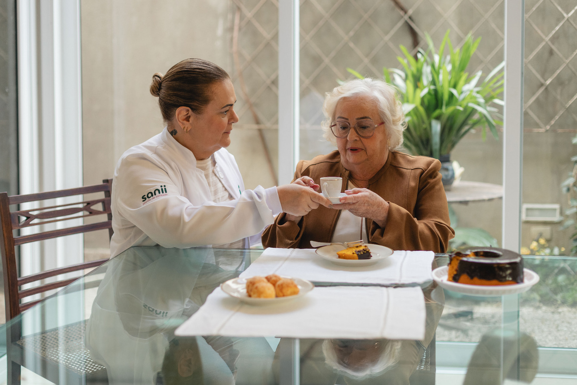 Cuidadora da Sanii servindo um café para uma senhora idosa à mesa, durante uma conversa tranquila
